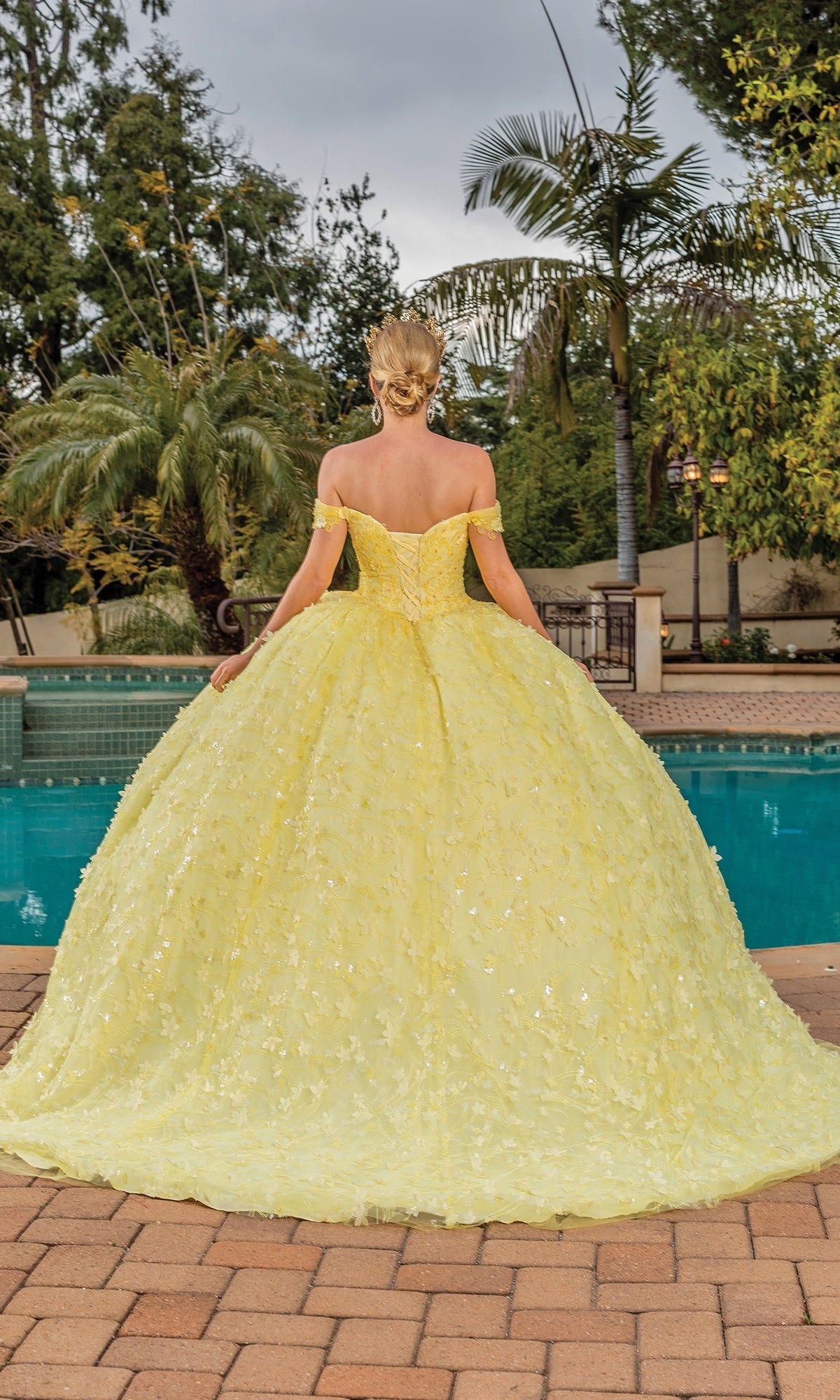 A person with blonde hair in the Quinceanera Dress 1757 by Dancing Queen, a yellow off-the-shoulder ball gown, stands on a stone patio looking at a swimming pool and lush green trees.